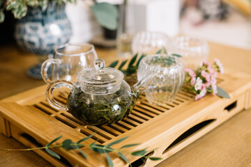 Delicious freshly brewed green tea ceremony. Glass tea cups with transparent glass tea pot standing on the wooden tray, decorated with small flowers 