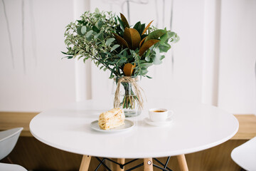 Delicious piece of cake an freshly brewed espresso coffee in a cup, standing ob the white table, decorated with transparent vase full of eucalyptus and magnolia