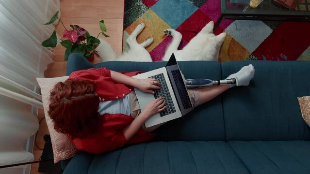 Top view happy disabled business woman with futuristic bionic leg prosthesis works online using laptop at home with dog.