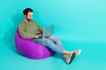 Full length profile side photo of young guy sit violet armchair use laptop chat report isolated over turquoise color background