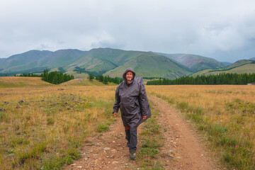 Fototapeta na wymiar Satisfied tourist returns from mountains in overcast. Happy man in raincoat walks through hills and forest in bad weather. Traveler goes towards adventure. Hiker and mountain range under cloudy sky.