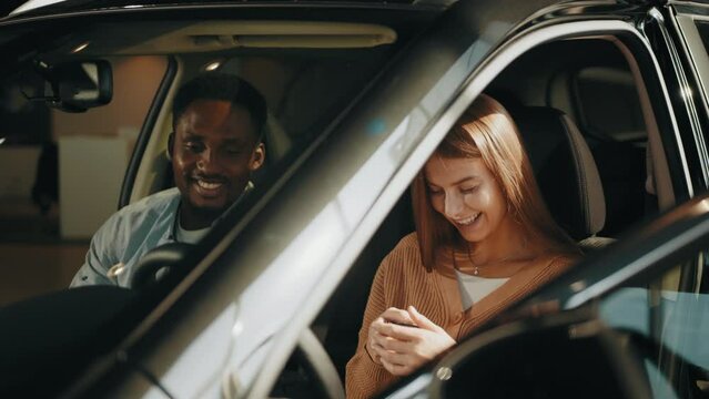 Salesman giving keys from new auto. Positive multi ethnic couple sitting inside luxury black car and talking with competent dealer at showroom. Man and woman choosing new vehicle
