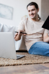 blurred man holding notebook and pen near laptop in bedroom.