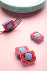 Dishwasher detergent tablet with fork and dishes on pink background. Vertical photo.