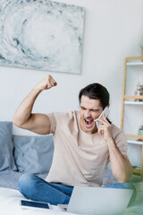 excited man talking on mobile phone near laptop in bedroom.