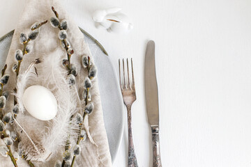 Stylish Easter table setting flat lay. Natural easter egg, pussy willow branches, feathers on modern plate with napkin and cutlery on white wooden table. Modern Easter table decoration