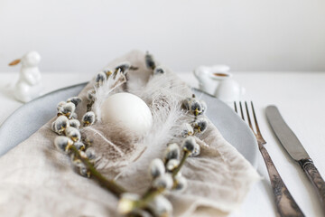 Stylish Easter table setting. Natural easter egg, pussy willow branches, feathers on modern plate with napkin and cutlery on white wooden table. Modern Easter table decoration