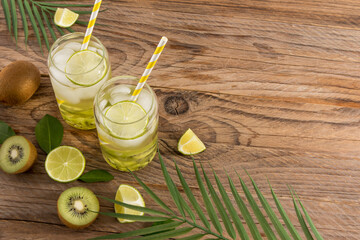 vitamin water with kiwi and lime in modern glasses in the form of a beer can against the background of a wooden table, fruits and tropical leaves.