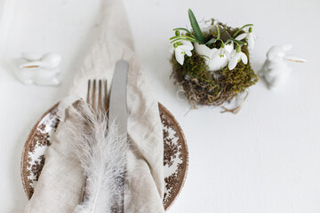 Stylish Easter table setting. Feathers on napkin on modern plate, cutlery, bunny and natural egg with snowdrops in nest on white wooden table. Modern Easter table rustic decoration