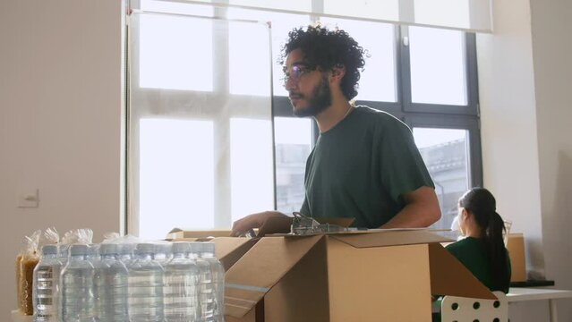 charity, donation and volunteering concept - male volunteers with clipboard packing food and drinks in boxes over international group of people at distribution or refugee assistance center