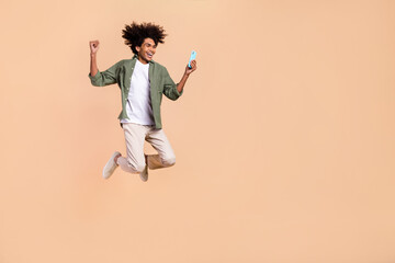 Fototapeta na wymiar Full length body size view of attractive cheerful trendy guy jumping using cell rejoicing isolated over beige pastel color background