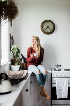 Vertical shot of a woman sitting on the kitchen counter near the window while drinking coffee