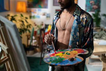 Attractive young man wearing unbuttoned shirt holding wooden pallete with oil paints and brush creating new artwork on canvas