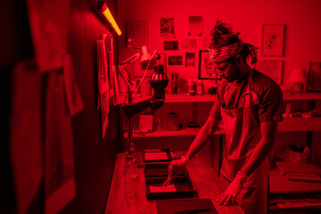 Creative young indian man wearing apron and protective gloves developing photos in red lit darkroom at home - obrazy, fototapety, plakaty