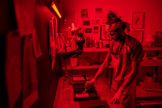 Creative young indian man wearing apron and protective gloves developing photos in red lit darkroom at home