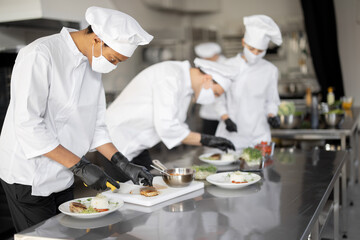 Fototapeta na wymiar Multiracial group of cooks finishing main courses while working together in the kitchen. Cooks wearing uniform and face mask. Team prepares meals for the restaurant