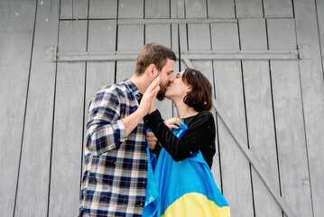 Kiss of husband and wife holding blue and yellow national flag of Ukraine on a background of gray wooden wall on the street in snowy weather. Support for the country during the occupation. Kyiv Day