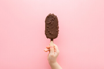 Baby girl hand holding ice cream with nuts and chocolate glaze on light pink table background....