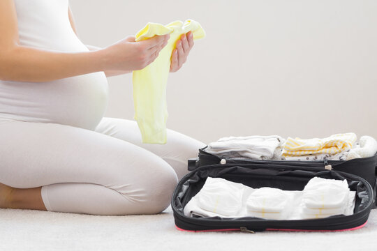 Young adult pregnant woman with big belly sitting on carpet and packing baby clothes and diapers in black bag. Hands holding bodysuit. Preparing things for hospital childbirth. Closeup. Side view.