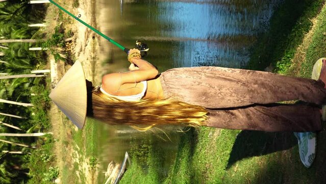 Vertical video, caucasian girl with long blonde hair wearing Asian cone hat, holding fishing pole, fishing on pond