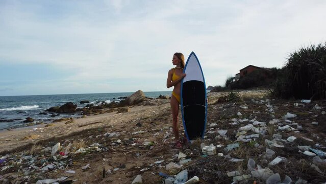 Beauty with yellow bikini and surfboard posing on beach covered in plastic garbage, orbit motion view