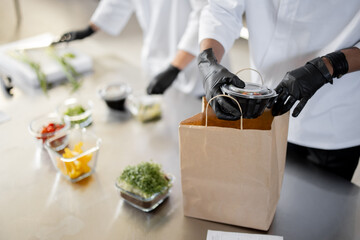 Cooks in protective gloves prepare take away food and packing them into paper bag for delivery,...