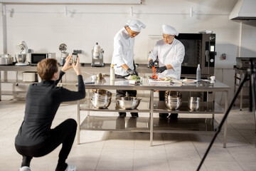 Man records on phone the process of cooking by two chefs in a professional kitchen. Latin-American and Asian cooks preparing food for social media. Concept of culinary video blogging