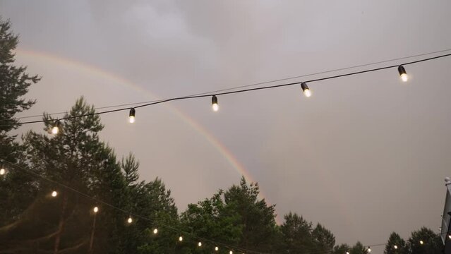 Bright beautiful rainbow in orm of arc over green forest in evening overcast sky with cloud on site with garland of glass bulbs of warm glow after rain. Smooth camera movement.