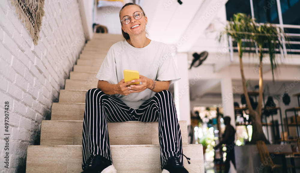 Poster portrait of cheerful hipster girl in optical spectacles smiling at camera during weekend pastime for