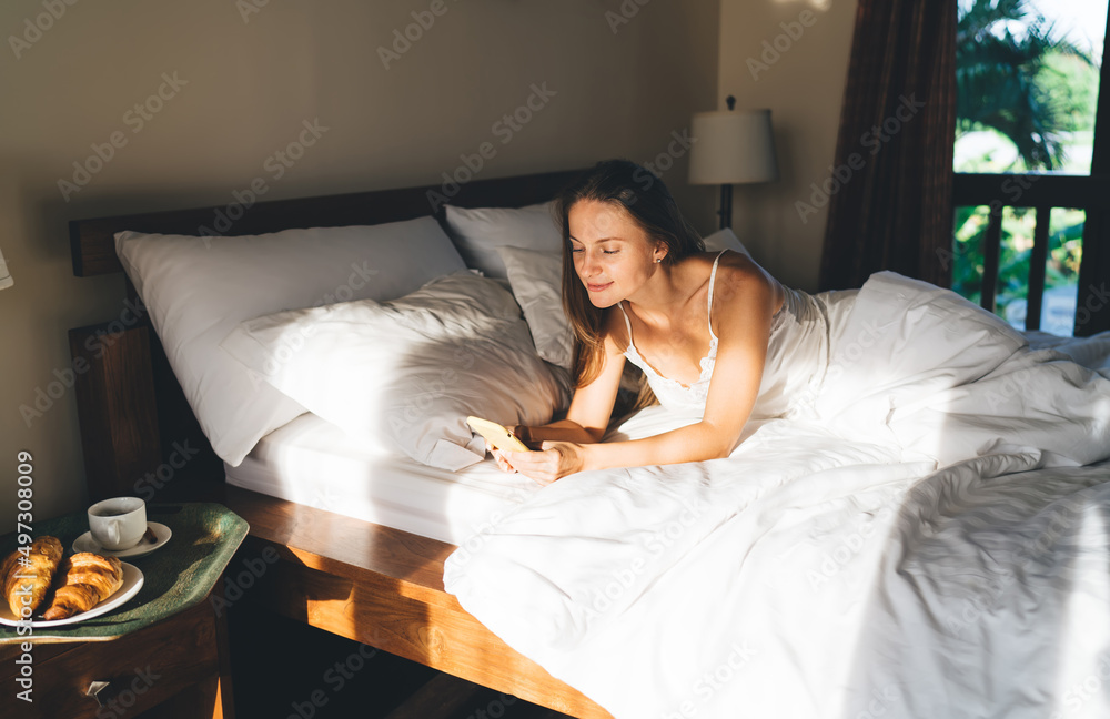 Poster caucaisan female blogger lying at comfortable bed with cellphone for browsing web page in networks, 