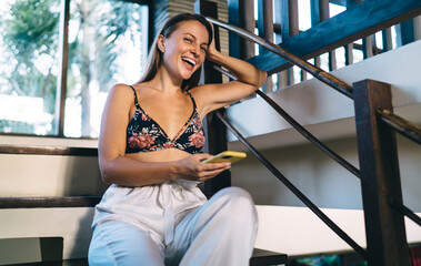 Portrait of millennial female blogger with digital mobile phone smiling at camera during pastime for online networking, cheerful Caucaisan woman with cellular gadget laughing while resting