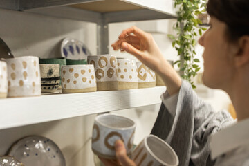 Young pretty woman artisan putting her ceramic works on exposition in her workshop