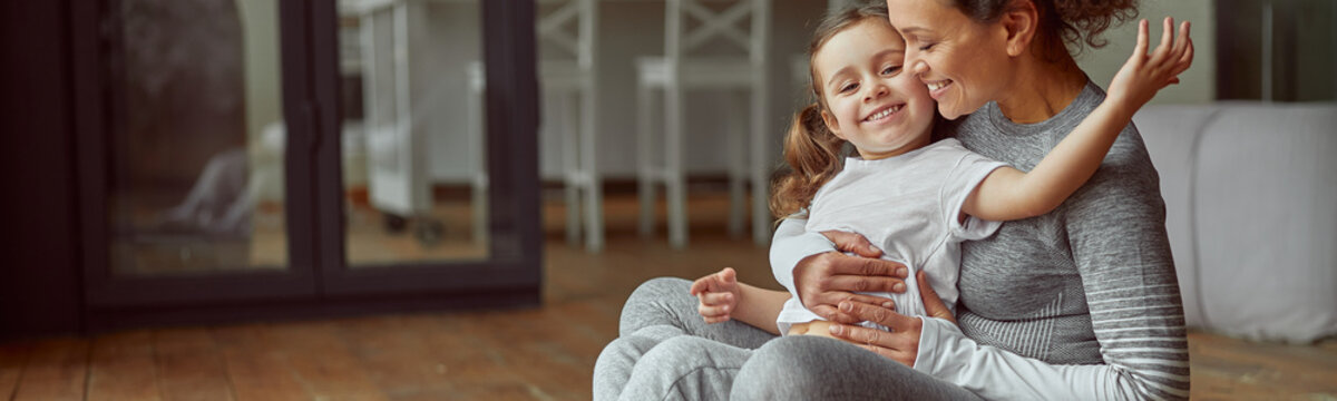 Jolly mother is hugging with little girl while doing workout together in cozy living room