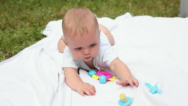 Little newborn girl lying on tummy chewing rattle on blanket, summer day outdoor. Infant having fun outdoors. Infant baby child resting playing learning to crawl. Motherhood teething, babyhood concept