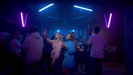 Two girls with a cocktail in their hands are dancing, a crowd of people is dancing around the girls on the dance floor of a nightclub under the light of colored spotlights. Girls take selfies on the