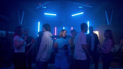 A pretty girl with a cocktail and a phone in her hands is dancing, a crowd of people are dancing around the girl on the dance floor of a nightclub under the light of colored spotlights. Night club