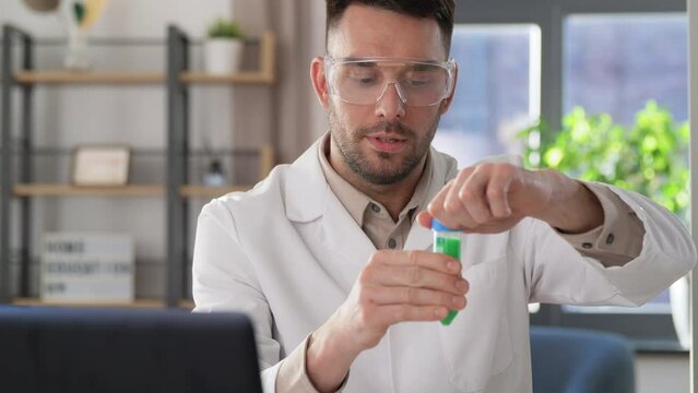 distance education, school and science concept - male chemistry teacher in goggles with test tubes making chemical reaction and having online class at home office