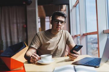 Handsome man in red casual t-shirt writing a message on mobile phone while sitting near glass...