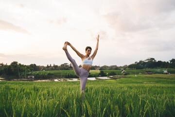 Flexible female in tracksuit stretching enjoying morning time for yoga practice, Caucasian woman in casual sportswear raising leg have hatha training for feeling vitality at rice fields in Vietnam