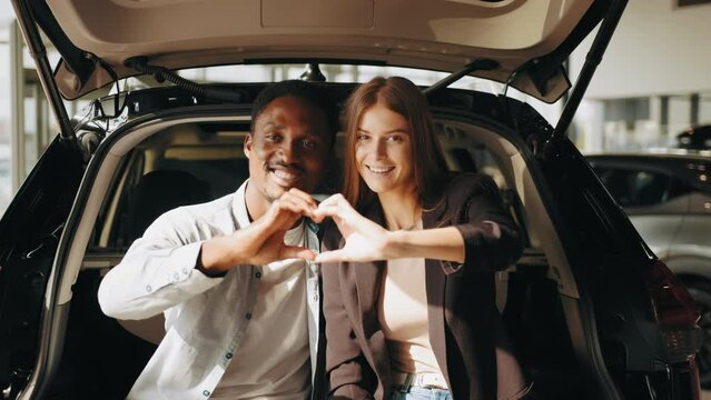 Smiling multi ethnic couple making heart shape with fingers while posing on camera in open trunk of their new car. Concept of people, love and expensive purchase. Couple making heart with fingers.