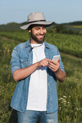 happy farmer in brim hat and denim shirt messaging on smartphone in field.