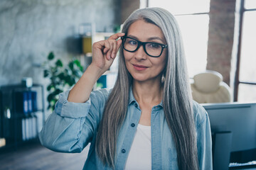 Portrait of charming satisfied aged lady hand touch eyeglasses look camera daylight workstation indoors