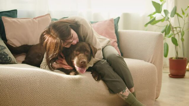 Brown dog close-up. Woman hugging and kissing retriever on sofa in living room, obedient labrador posing. Happy domestic animal concept, best friends, puppy relaxing at home, breathing with tongue out