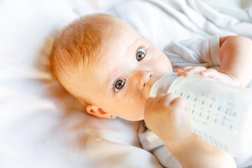 Cute little newborn girl drinking milk from bottle and looking at camera on white background....