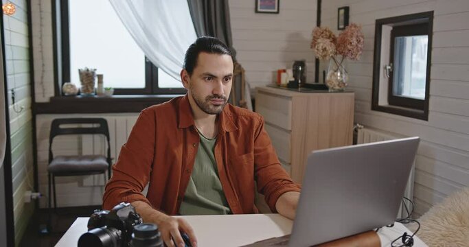 A young, handsome male photographer and videographer works at home on a laptop.