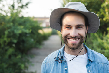 Naklejka na ściany i meble young and bearded farmer in brim hat smiling at camera outdoors.