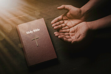A bible on a wooden table illuminated from above.  and praying hands  concept of faith, hope, trust...