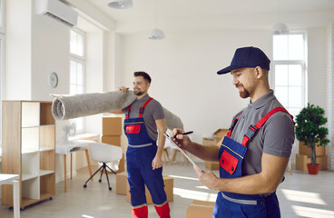 Obraz premium Two workers in uniforms remove things from old apartment. Happy man holding clipboard and taking notes while his teammate is carrying rugs and other stuff. Moving house, using delivery service concept