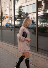 shopping walk leisure. glamour blonde girl in beige oversize dress is stands casual in step near street showcase store background and looks apart with black bag in hand. lifestyle concept, free space