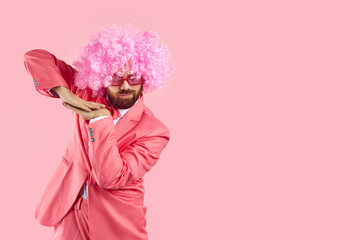 Funny goofy bearded adult man wearing pink party suit, curly clown wig and funky sunglasses dancing...